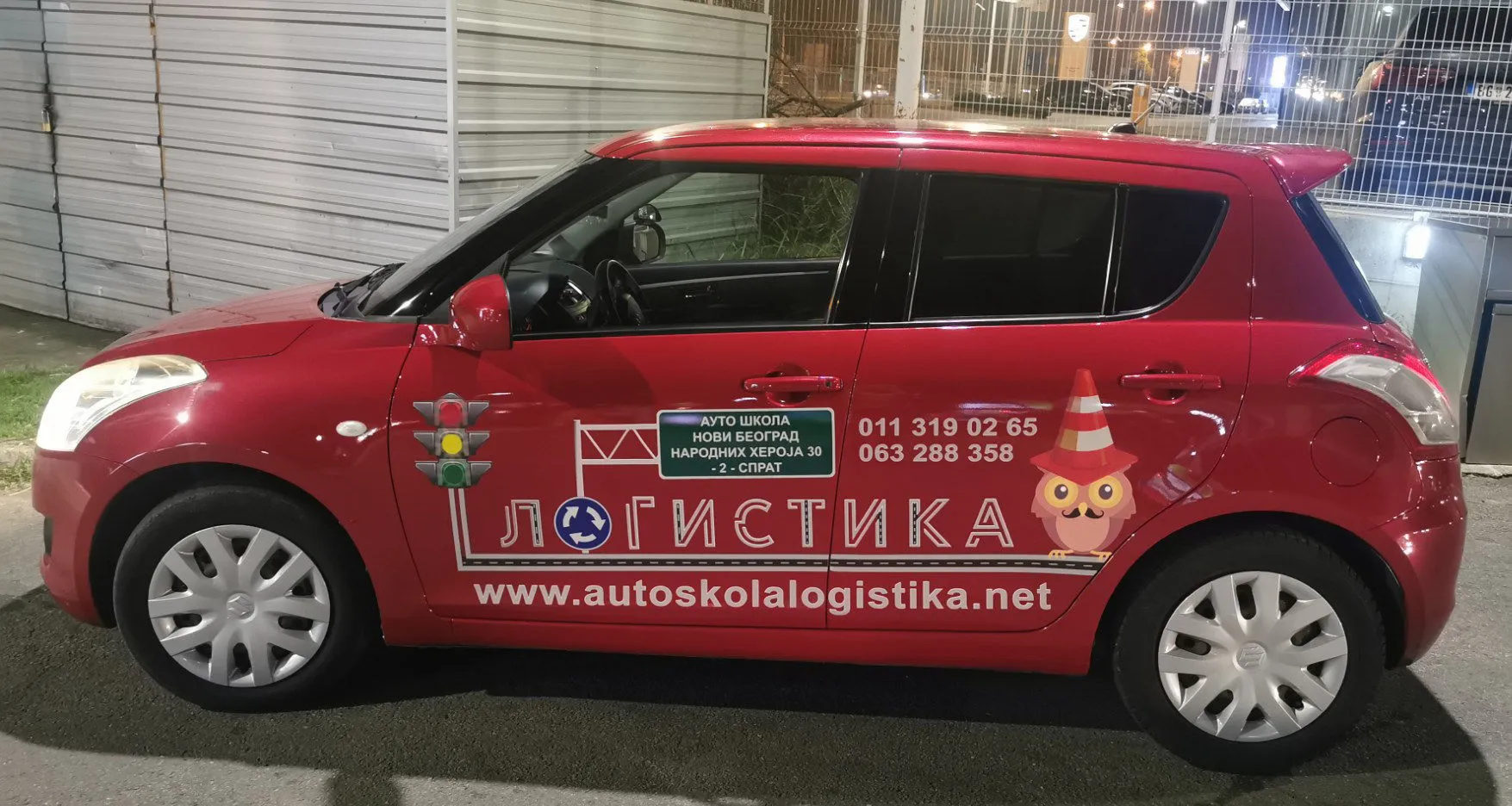 Vozilo auto škole Logistika sa nalepnicom loga, maskote sove i brojeva telefona, parkirano napolju.